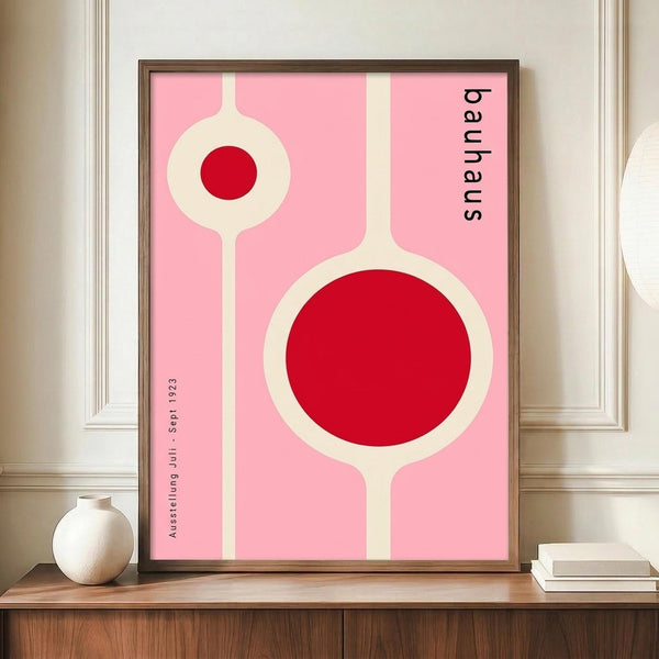 May include: Framed Bauhaus-style poster with pink background, white geometric circular forms, and red circles, displayed on wooden sideboard next to white ceramic vase and books in elegant room with cream walls and crown molding.
