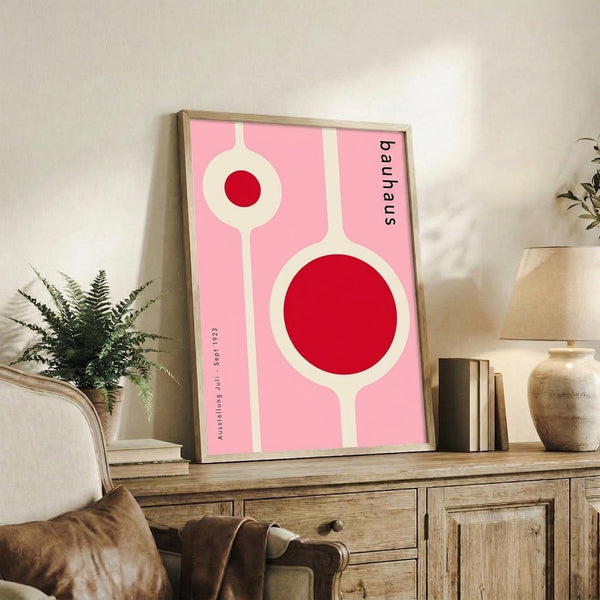 May include: Pink and red geometric Bauhaus-style poster in thin wooden frame, displayed on rustic wood sideboard alongside fern plant, books, and ceramic vase. The minimalist artwork features bold circular shapes and clean lines against a soft pink background. Cozy living room setting with beige upholstered furniture and neutral wall.