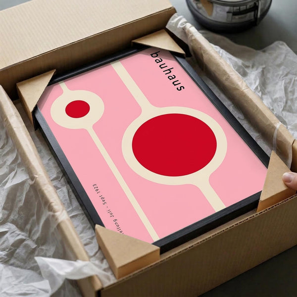 May include: Pink and red Bauhaus-style poster with geometric circular shapes in black frame, partially unwrapped from cardboard packaging with tissue paper, surrounded by craft supplies and wooden handles on neutral surface