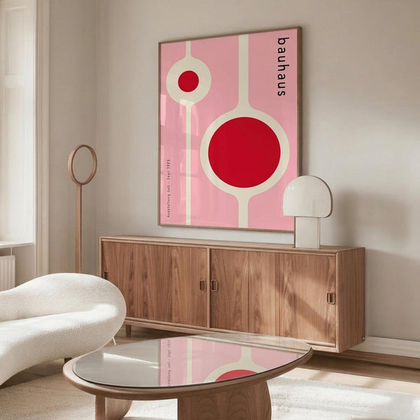 May include: Pink Bauhaus exhibition poster with red circles and white geometric shapes mounted on white wall above wooden mid-century sideboard. Modern living room setting with cream sculptural chair, glass coffee table, and white table lamp. Poster features vintage typography and minimalist design in soft pink tones.