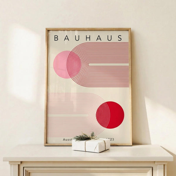 May include: A framed Bauhaus-style poster with geometric pink and red circular shapes intersected by curved parallel lines, displayed on a light wood console table beside a small wrapped gift with twine and evergreen sprig. The minimalist artwork features a cream background with the word "BAUHAUS" at the top, presented in a natural wood frame against a white wall.