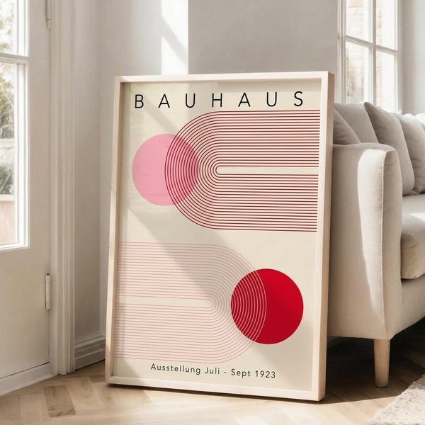 May include: Framed Bauhaus-style poster with geometric design featuring pink and red circles intersecting with curved linear patterns on cream background, displayed against white wall next to beige upholstered chair in bright living room with large windows and natural lighting