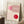 May include: Framed Bauhaus exhibition poster with geometric red and pink circular designs and curved line patterns, displayed in a light wooden frame leaning against white paneled walls. A white coffee cup sits on a wooden block nearby, with a cream textured rug and beige blanket visible in the cozy interior setting.