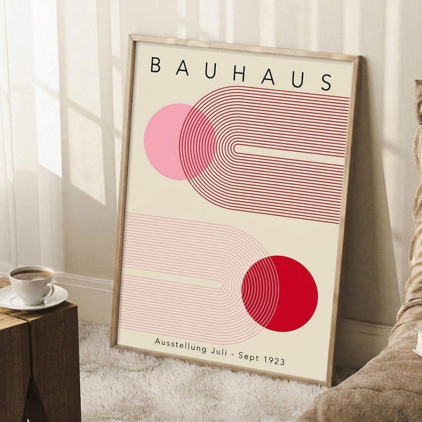 May include: Framed Bauhaus exhibition poster with geometric red and pink circular designs and curved line patterns, displayed in a light wooden frame leaning against white paneled walls. A white coffee cup sits on a wooden block nearby, with a cream textured rug and beige blanket visible in the cozy interior setting.