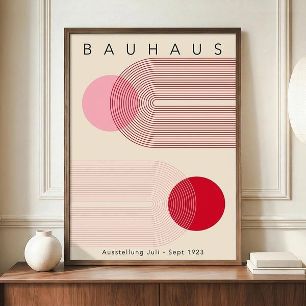 May include: Framed Bauhaus exhibition poster in modern minimalist style featuring geometric red and pink circular forms with curved linear patterns on cream background, displayed on wooden console beside white ceramic vase and books in elegant room with cream walls and architectural molding.