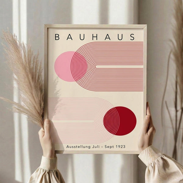 May include: Person holding framed Bauhaus exhibition poster with geometric design featuring pink and red curved lines and circles on cream background. Dried pampas grass visible to left. Neutral home interior with beige clothing and soft lighting.