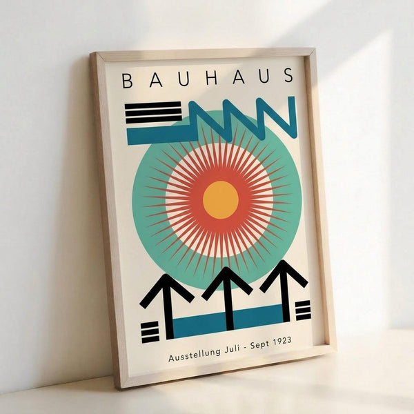 Framed poster with cream background featuring "BAUHAUS" text at top, geometric design with large teal circle containing radiating red and white sunburst pattern around yellow center, black angular shapes resembling building rooftops at bottom, blue horizontal bands, and "Ausstellung Juli - Sept 1923" text below, displayed in light wood frame leaning against white wall