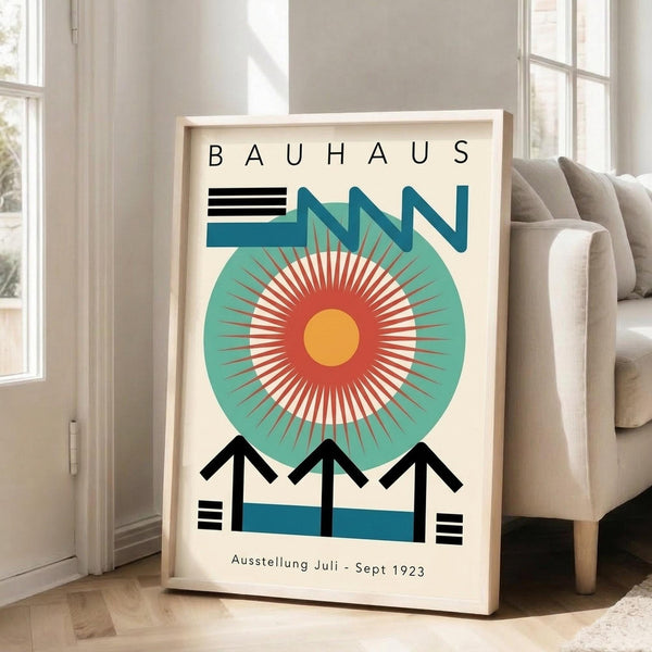 May include: Framed Bauhaus-style poster with geometric design featuring large teal circle, radiating orange and red sunburst pattern, blue zigzag elements, and black angular shapes. Light wood frame leaning against white wall next to beige sofa in bright room with large windows and natural lighting.