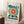 May include: Framed Bauhaus-style poster leaning against white wall featuring geometric design with large teal circle, radiating orange and red sunburst pattern, black angular shapes, and "BAUHAUS" text at top. Cream-colored background with wooden frame. Scene includes white coffee cup on wooden block and textured rug in cozy interior setting with natural lighting.
