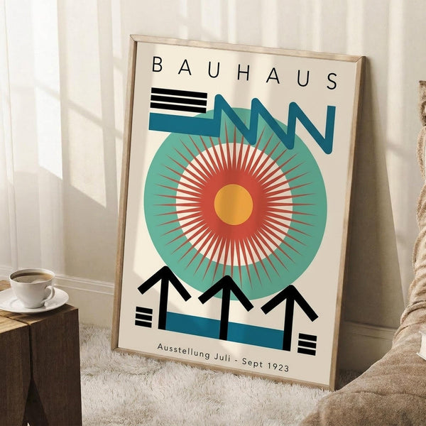 May include: Framed Bauhaus-style poster leaning against white wall featuring geometric design with large teal circle, radiating orange and red sunburst pattern, black angular shapes, and "BAUHAUS" text at top. Cream-colored background with wooden frame. Scene includes white coffee cup on wooden block and textured rug in cozy interior setting with natural lighting.