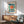 May include: Bauhaus exhibition poster in wooden frame hanging above mid-century modern furniture. The geometric design features a large teal circle with radiating orange and red sunburst pattern, black angular shapes below, and "BAUHAUS" text at top. Room shows light wood sideboard, sage green upholstered chair with wooden arms, white ceramic lamp, and natural lighting casting shadows on white wall.
