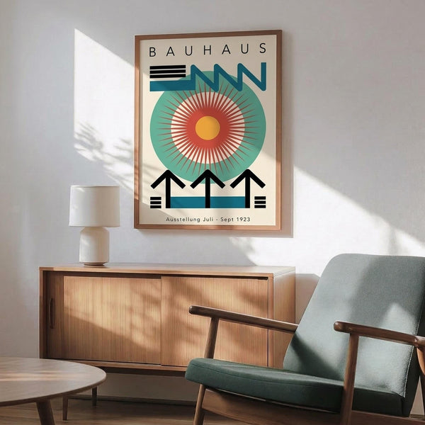 May include: Bauhaus exhibition poster in wooden frame hanging above mid-century modern furniture. The geometric design features a large teal circle with radiating orange and red sunburst pattern, black angular shapes below, and "BAUHAUS" text at top. Room shows light wood sideboard, sage green upholstered chair with wooden arms, white ceramic lamp, and natural lighting casting shadows on white wall.