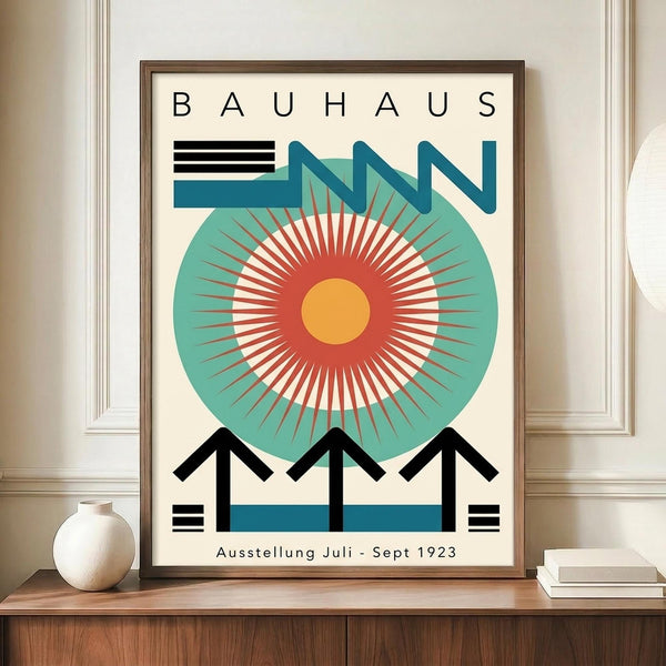 May include: Framed Bauhaus-style poster with geometric design featuring a large circular sunburst in coral, yellow and teal colors, black angular shapes, and "BAUHAUS" text at top. The cream-colored print is displayed in a brown wooden frame on a mid-century modern wooden sideboard next to a white ceramic vase and stack of books in a minimalist room setting.