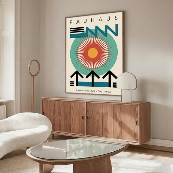 May include: Vintage Bauhaus exhibition poster with geometric design featuring large teal circle, radiating orange and red sunburst pattern, black angular shapes, and "BAUHAUS" text at top. Displayed in thin brown frame above walnut credenza in modern living room with cream curved chair, glass oval coffee table, and white table lamp.