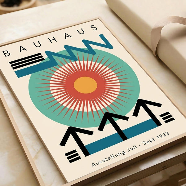 May include: Vintage Bauhaus exhibition poster from 1923 displayed in cream wooden frame on white surface. Features bold geometric design with circular radiating sunburst pattern in red, orange and teal, black angular text elements, and "Ausstellung Juli - Sept 1923" at bottom. Modernist art print with clean lines and vibrant retro color palette.