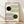 May include: Framed Bauhaus poster print leaning against white wall on wooden floor. Cream background with geometric design featuring black and green circles intersected by flowing curved parallel lines. Modern minimalist aesthetic with clean typography reading "BAUHAUS" and "Ausstellung Juli - Sept 1923". Natural wood frame, soft window lighting creating diagonal shadows on wall.