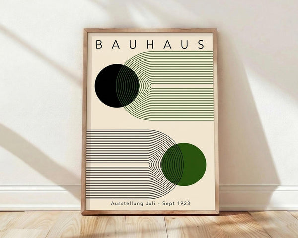 May include: Framed Bauhaus poster print leaning against white wall on wooden floor. Cream background with geometric design featuring black and green circles intersected by flowing curved parallel lines. Modern minimalist aesthetic with clean typography reading "BAUHAUS" and "Ausstellung Juli - Sept 1923". Natural wood frame, soft window lighting creating diagonal shadows on wall.