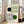 May include: Framed Bauhaus-style minimalist poster with geometric line art featuring black and green circles with curved linear patterns, displayed in light wooden frame leaning against white wall next to beige upholstered chair in bright, airy living room with large windows and natural lighting.