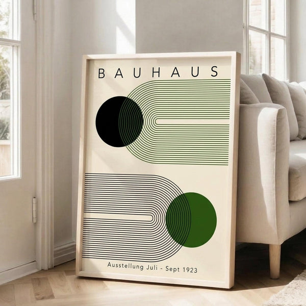 May include: Framed Bauhaus-style minimalist poster with geometric line art featuring black and green circles with curved linear patterns, displayed in light wooden frame leaning against white wall next to beige upholstered chair in bright, airy living room with large windows and natural lighting.