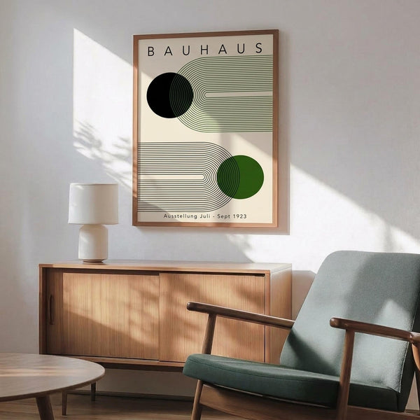 May include: Framed Bauhaus exhibition poster with geometric design featuring black and green circles connected by curved linear patterns on cream background, displayed above mid-century modern wood credenza with ceramic lamp, next to sage green upholstered armchair with wooden arms in sunlit room with white walls