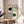 May include: Bauhaus-style poster print in wooden frame displayed on round wooden stool next to beige upholstered chair. Design features geometric black and green circles with curved concentric lines forming abstract shapes on cream background. Soft natural lighting creates shadows from nearby plant leaves on neutral wall.
