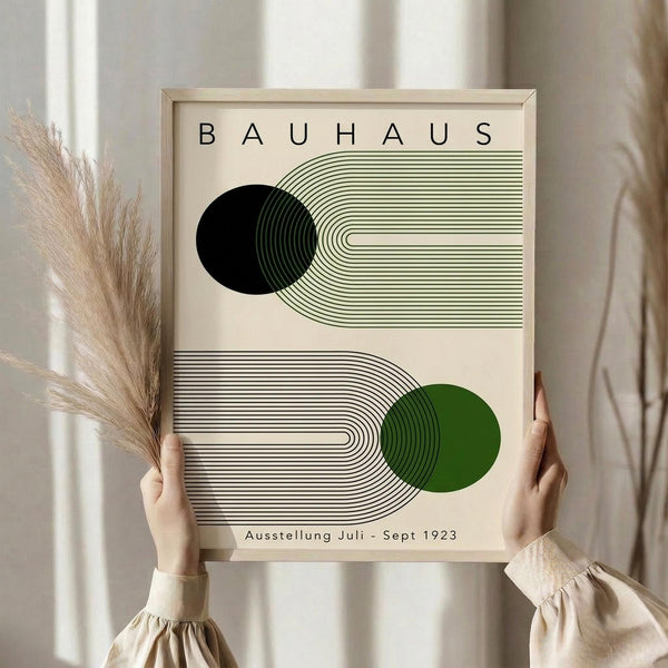 May include: Person holding a framed Bauhaus-style poster featuring geometric line art with black and green circles intersecting curved linear patterns. The minimalist design has a cream background with "BAUHAUS" text at top and "Ausstellung Juli - Sept 1923" at bottom. Light wood frame held by hands in beige sleeves, with dried pampas grass visible to the left against a neutral curtained background.