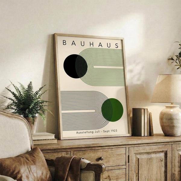 May include: Framed Bauhaus-style poster with geometric design featuring black and green circles intersected by curved linear patterns, displayed on rustic wooden sideboard alongside fern plant, ceramic vase, and leather armchair in modern neutral-toned living room setting.