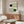 May include: Modern living room displaying framed Bauhaus exhibition poster on beige wall above wooden sideboard. Poster features geometric design with black and green circles, curved linear patterns on cream background. Room styled with white curved chair, glass oval coffee table, contemporary lamp, and neutral decor creating minimalist aesthetic.