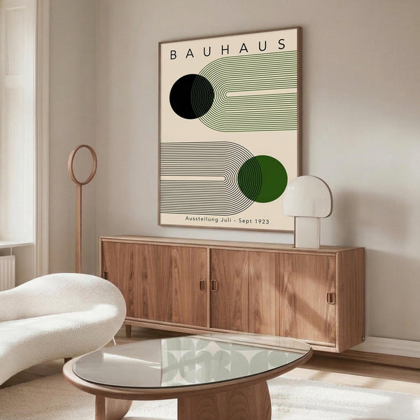 May include: Modern living room displaying framed Bauhaus exhibition poster on beige wall above wooden sideboard. Poster features geometric design with black and green circles, curved linear patterns on cream background. Room styled with white curved chair, glass oval coffee table, contemporary lamp, and neutral decor creating minimalist aesthetic.