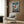 May include: One Piece anime poster mounted on wood-paneled wall above beige armchair with geometric pillow. Artwork shows Luffy in red shirt and straw hat leaping from small boat against stormy ocean backdrop with Japanese text. Modern living room setting features brass floor lamp, dark round side table with open book, and neutral tones throughout.