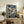 May include: Framed One Piece anime poster featuring Luffy in red shirt and straw hat leaping from wooden boat over stormy ocean waters with dramatic cloudy sky, displayed on rustic wooden sideboard next to fern plant, pottery vase, and leather armchair in cozy living room setting with neutral beige walls