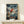 May include: Framed One Piece anime poster showing Luffy in his signature red vest and straw hat leaping from a wooden sailing boat across choppy ocean waters under dramatic stormy skies. The artwork features vibrant colors with Japanese text and "Journey Beyond the Horizon" subtitle. Light wood frame sits on a minimalist white shelf next to a small white wrapped gift against a cream wall.