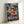May include: Framed anime poster showing character with straw hat throwing flaming punch on stormy ship deck. Lightning strikes dark sky behind sailing vessel with rigging and masts. Character wears red vest, determined expression. Japanese text overlays dramatic blue and orange scene. Light wooden frame leans against white wall.