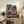 May include: Framed anime poster displayed on rustic wooden sideboard in cozy living room setting. Artwork shows character in red outfit and straw hat with flaming fist on stormy ship deck, featuring blue lightning effects and Japanese text. Frame has light wood finish, surrounded by fern plant, ceramic vases, books, and cream upholstered furniture.