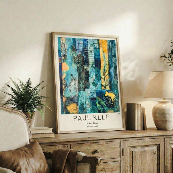 May include: Framed abstract art print displayed on rustic wooden sideboard in modern living room setting. Artwork features vibrant blue and teal botanical motifs with orange accents, geometric patterns, and leaf designs. Light wood frame against white wall, styled with fern plant, ceramic vases, books, and beige upholstered chair visible in foreground.