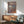 May include: Japanese ukiyo-e style artwork in wooden frame showing geisha in colorful kimono seated on wooden platform with snow-capped Mount Fuji and traditional buildings in background, displayed above mid-century modern walnut credenza with white ceramic lamp, next to sage green upholstered armchair with wooden arms in bright sunlit room with white walls
