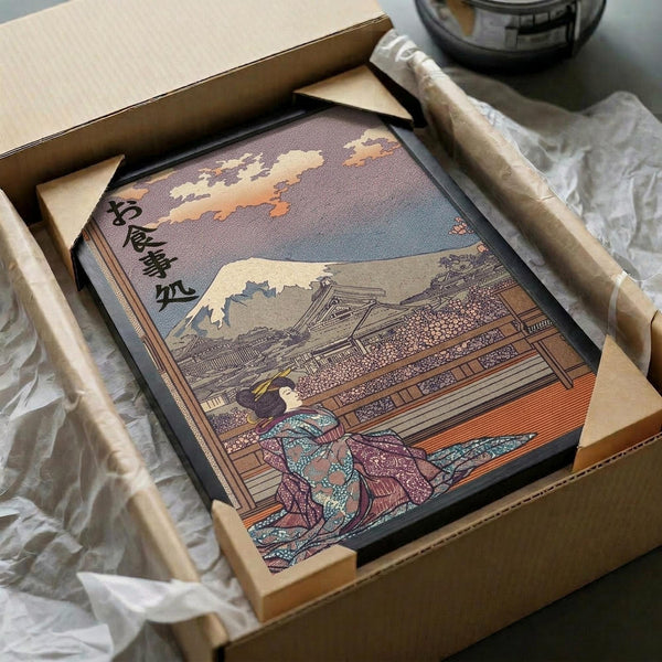 Traditional Japanese woodblock print artwork in ornate black frame, featuring a geisha in colorful robes sitting beside a wooden building with Mount Fuji and cloudy sky in the background. The print shows intricate details in earth tones, blues, and warm colors with Japanese calligraphy in the upper left corner. Packaged in brown cardboard box with protective wrapping and wooden dowel rods, photographed on white tissue paper.