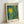 A framed poster featuring abstract art with a bright yellow sun surrounded by radiating vertical streaks in teal, green, and yellow tones, dotted with circular polka dot patterns. The artwork has "YAYOI KUSAMA" at the top, "Modern Art" at the bottom left, and "1964" at the bottom right on a cream background. The natural wood frame leans against a white wall, casting a soft shadow.