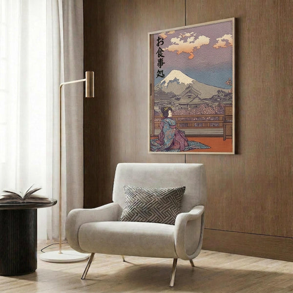May include: Japanese ukiyo-e style artwork displayed in modern living room setting featuring geisha figure with Mount Fuji backdrop and Japanese text, warm sunset colors, framed in light wood, hung above beige upholstered chair with geometric pillow, brass floor lamp and dark round side table with open book nearby, wood panel wall background.