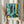 May include: Person in cream ruffled sleeves holding framed Paul Klee poster against white curtained window. Abstract artwork features vibrant blues, teals, oranges and yellows with layered botanical motifs, geometric patterns, and fish silhouettes. Dried pampas grass visible on left. Light wooden frame with white matting displays "La Mer Bleue Switzerland" text below colorful collage-style composition mixing organic and architectural elements.