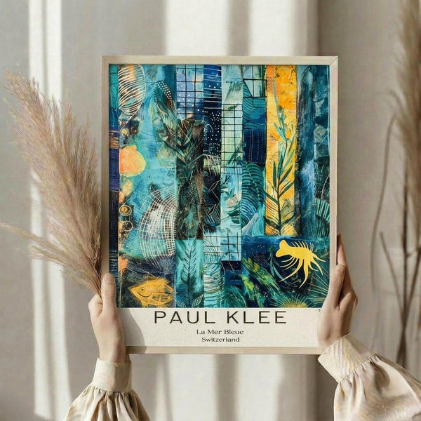 May include: Person in cream ruffled sleeves holding framed Paul Klee poster against white curtained window. Abstract artwork features vibrant blues, teals, oranges and yellows with layered botanical motifs, geometric patterns, and fish silhouettes. Dried pampas grass visible on left. Light wooden frame with white matting displays "La Mer Bleue Switzerland" text below colorful collage-style composition mixing organic and architectural elements.