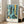 May include: Framed art print leaning against white wall next to beige sofa in bright living room. Abstract artwork features blue and teal underwater scene with geometric patterns, coral-like forms, yellow fish, and tropical motifs. White border displays "PAUL KLEE" and "La Mer Bleue Switzerland 1924" text. Silver frame sits on light wooden floor with natural window light creating shadows on wall.