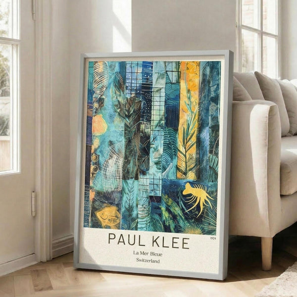 May include: Framed art print leaning against white wall next to beige sofa in bright living room. Abstract artwork features blue and teal underwater scene with geometric patterns, coral-like forms, yellow fish, and tropical motifs. White border displays "PAUL KLEE" and "La Mer Bleue Switzerland 1924" text. Silver frame sits on light wooden floor with natural window light creating shadows on wall.