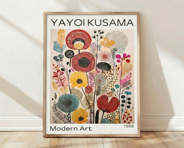 May include: Framed poster print leaning against white wall on light wood floor. Features colorful botanical illustration with stylized flowers, leaves and stems in red, yellow, teal, pink and purple on cream background. Text reads "YAYOI KUSAMA" at top and "Modern Art 1966" at bottom. Natural wood frame casting soft shadows on textured white wall.