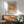 May include: Framed Mark Rothko-inspired abstract art poster with warm earth tones - pink, olive green, and cream color blocks - hanging above a mid-century modern wooden sideboard. Green upholstered armchair with wooden frame sits nearby, along with a small round coffee table and white ceramic table lamp. Soft natural lighting creates shadows on white wall in minimalist living room setting.