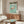 May include: Japanese flamingo exhibition poster in wooden frame hanging above mid-century walnut credenza in minimalist living room. Poster features pink flamingo on turquoise background with black Japanese text. White curved chair and oval glass coffee table with abstract art visible. Brass floor lamp and table lamp provide warm lighting against neutral walls.