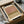 May include: Abstract art print with horizontal bands of muted pink, olive green, and beige tones in earthy color palette, displayed in black frame within brown cardboard packaging box surrounded by white tissue paper on neutral surface