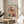 May include: Framed art print featuring colorful stylized flowers and botanical elements in red, yellow, blue, and pink tones displayed on a round wooden side table next to a beige upholstered chair. Natural sunlight creates soft shadows on white walls. The artwork shows decorative floral motifs with geometric patterns and is presented in a light wood frame.