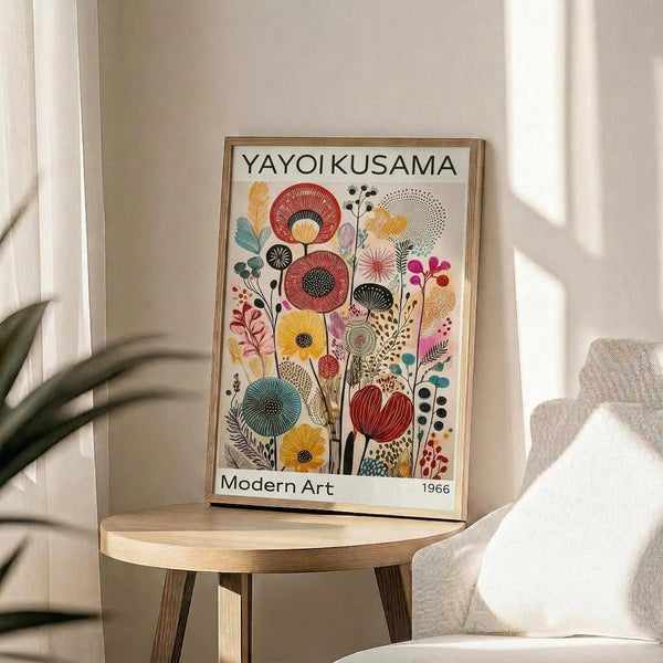 May include: Framed art print featuring colorful stylized flowers and botanical elements in red, yellow, blue, and pink tones displayed on a round wooden side table next to a beige upholstered chair. Natural sunlight creates soft shadows on white walls. The artwork shows decorative floral motifs with geometric patterns and is presented in a light wood frame.