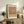 May include: Framed art print with abstract horizontal color blocks in muted pink, olive green, and cream tones, displayed on rustic wooden sideboard alongside potted fern, ceramic vase, and stacked books in neutral-toned living room setting with white walls and upholstered furniture.